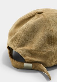 Casquette en daim marron avec une forme arrondie, présentant une surface texturée, une sangle à l'arrière avec une quincaillerie en métal, et un détail en bouton sur le dessus.