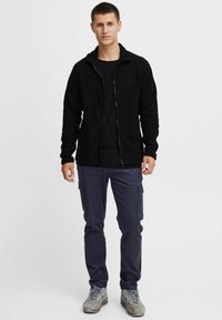 Sort fleece zip-up jakke, tætsiddende snit, lange ærmer. Båret over en sort skjorte med mørke cargos og grå sportsko.