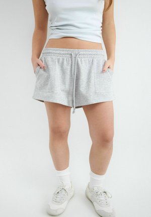 Femme portant un short gris clair à cordon de serrage avec poches avant, des baskets blanches, des chaussettes blanches et un débardeur blanc court, debout les mains dans les poches.