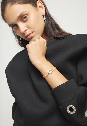 Mujer con blusa negra de mangas abullonadas que muestra una pulsera de cordón naranja con un dije de corazón de plata y pendientes a juego de plata.