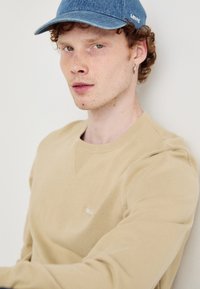 Écharpe beige avec texture côtelée, avec une petite étiquette logo sur la poitrine. Casquette en denim avec logo, cheveux bouclés visibles. Fond neutre.