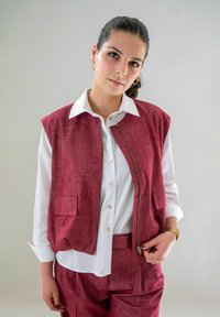 Vrouw met donker haar in een paardenstaart draagt een witte blouse, een bordeauxrood textuurvest en bijpassende broek, staand tegen een neutrale achtergrond.