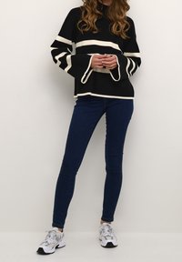 Czarnobiały prążkowany sweter z rozkloszowanymi rękawami, zestawiony z ciemnoniebieskimi dopasowanymi jeansami i szarymi butami sportowymi.