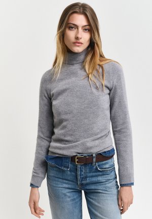 Junge Frau mit langen blonden Haaren, die einen grauen Rollkragenpullover, ein blaues Jeanshemd darunter, blaue Jeans und einen braunen Gürtel trägt.