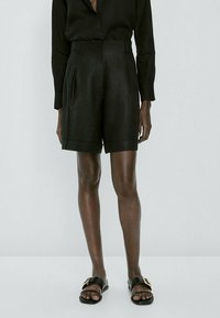 Personne portant un short noir taille haute sur mesure, un haut noir à manches longues et des sandales noires avec des boucles dorées, debout sur un fond clair.