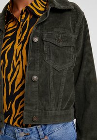 Ciemnozielona kurtka z kordurowego materiału z kołnierzem, z dwoma przednimi kieszeniami i srebrnymi guzikami, noszona na wzorzystą żółtą koszulę.