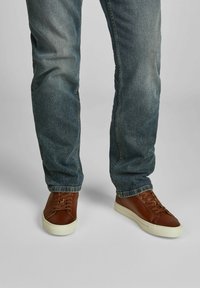 Beine in verblassten blauen Jeans und braunen Leder-Sneakern mit weißen Sohlen stehen vor einem grauen Hintergrund.