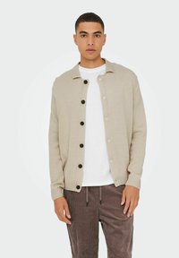 Beige strikket cardigan med knaplukning foran, ribbede manchetter og krave. Båret over en hvid T-shirt og brune kordfløjlsbukser.