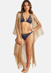 Gouden haakwerk cover-up met franjes, gedragen over een marineblauwe bikini met decoratieve borduring en een driehoekige top.