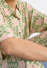 Chemise à col vert clair avec un motif ondulé. Présente des accents floraux roses, des manches courtes et des boutons bruns. Tissu doux et texturé.