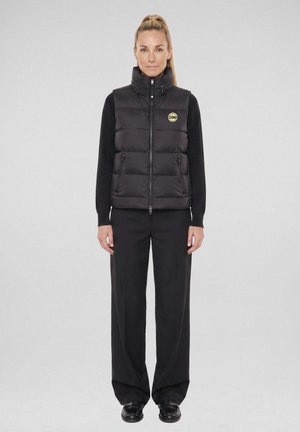 Vrouw staand met een zwart puffervest met een geel rond logo, een zwart shirt met lange mouwen, brede zwarte broek en zwarte schoenen tegen een grijze achtergrond.
