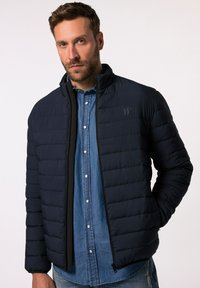 Giacca puffer blu marino con design trapuntato, colletto alto, zip nera e logo sottile sul petto, abbinata a una camicia di denim.