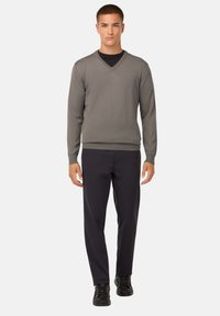 Grauer V-Ausschnitt-Pullover aus strukturiertem Stoff, kombiniert mit schwarzen Hosen und schwarzen Sneakers. Das Modell steht aufrecht mit den Händen seitlich.