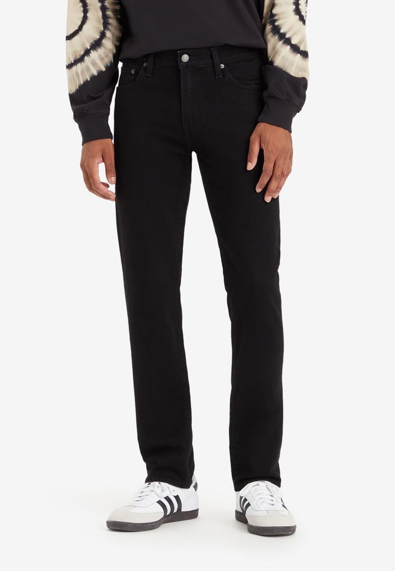 Homme portant un jean slim noir, des baskets blanches avec des bandes noires, et une chemise noire à manches longues avec des manches tie-dye beige, debout.