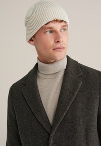 Cappello a maglia in avorio, con una texture a coste. Il modello indossa un blazer in tweed marrone sopra un maglione a collo alto grigio chiaro.