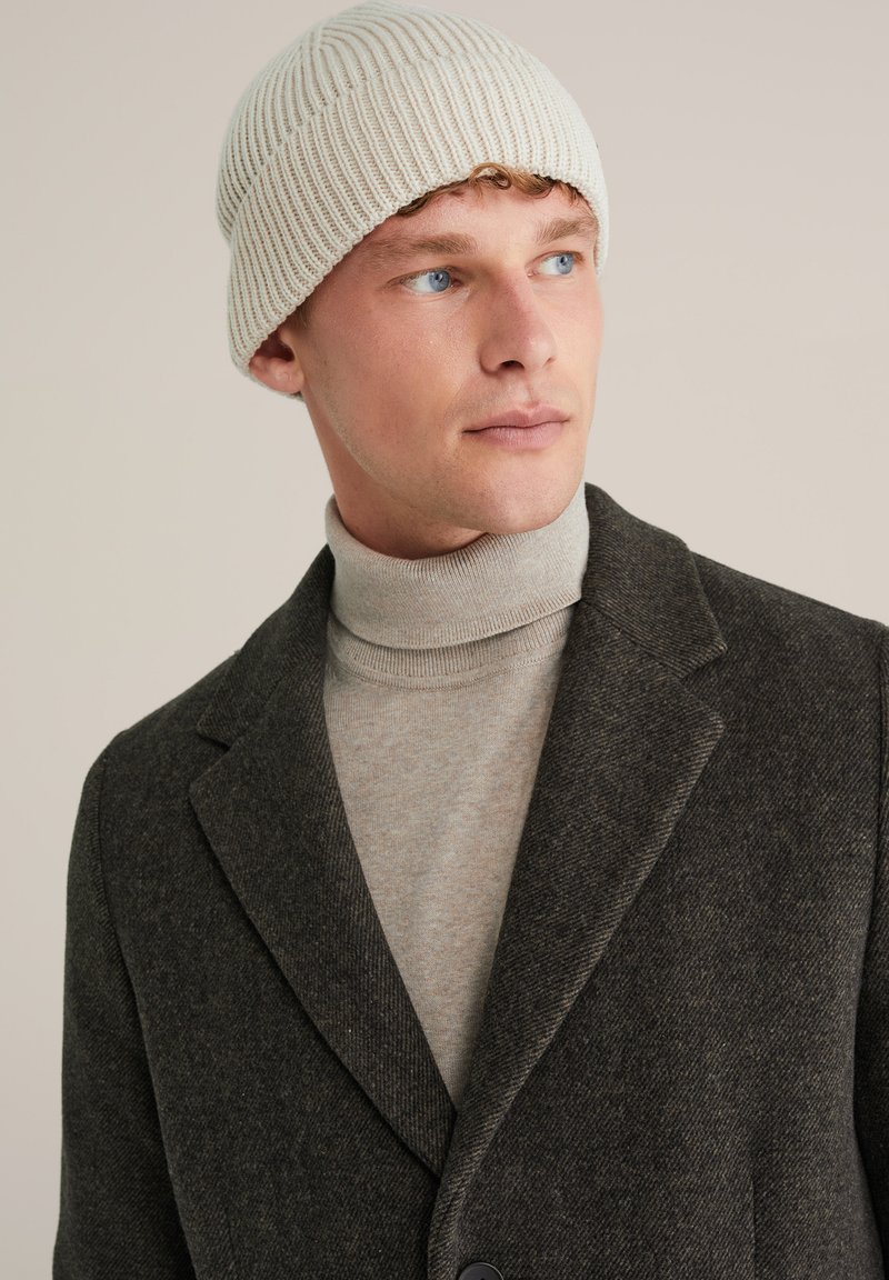 Cappello a maglia in avorio, con una texture a coste. Il modello indossa un blazer in tweed marrone sopra un maglione a collo alto grigio chiaro.