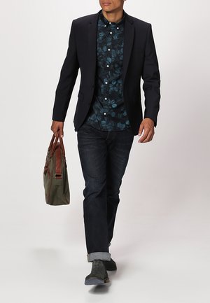 Hombre caminando con blazer negro, camisa de botones con estampado floral, jeans oscuros con dobladillos, zapatos grises, y llevando un bolso tote de cuero verde y marrón.