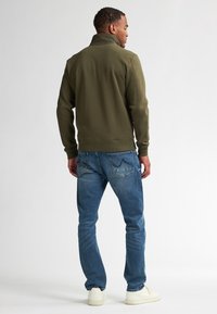 Olijfgroene ritsjack met hoge kraag, gecombineerd met blauwe jeans en witte sneakers, gezien vanaf de achterkant. Glad materiaal, casual ontwerp.