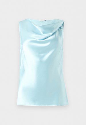 Kvinde iført lyseblåt satin top uden ærmer og matchende midi nederdel, stående med én hånd bag ryggen og et neutralt udtryk.