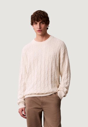 Jonge man met krullend haar die een gestructureerde off-white trui en lichtbruine broek draagt, staand tegen een effen lichte achtergrond.