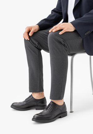 Mann sitzt auf einem Stuhl, trägt dunkelgraue Hosen, schwarze Brogue-Schnürschuhe und ein dunkelblaues Sakko, die Hände ruhen auf den Knien.