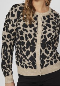 Cardigan beige à imprimé léopard noir, poignets et ourlet côtelés, avec une fermeture à boutons sur le devant. Tissu en maille texturée.