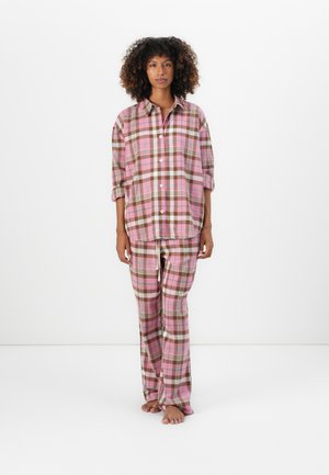 Femme debout, pieds nus, portant une chemise et un pantalon à carreaux rose et marron assortis, devant un fond blanc.