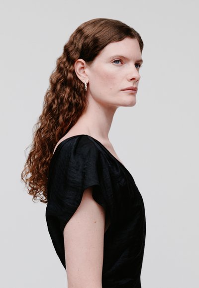 Γυναίκα με μακριά σγουρά καστανά μαλλιά, φοράει μαύρο τοπ με κοντά μανίκια και ασημένια σκουλαρίκια κρίκους, κοιτάζει προς τα δεξιά σε ανοιχτό φόντο.