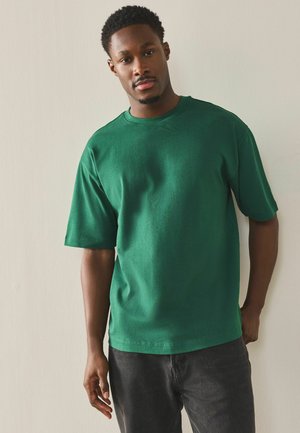 Young man with short curly hair wearing a green short-sleeve shirt and dark pants, standing against a plain light wall.