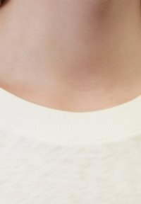 Vit bomullsplagg med ribbad rund hals, slät textur och subtila sömnadsdetaljer runt kragen.