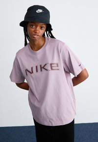 Camiseta de algodón lavanda con un gran gráfico de "NIKE" en burdeos oscuro. Combinada con un sombrero tipo bucket negro que presenta un logo blanco, el modelo posa frente a un fondo liso.