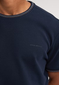 Námořnická modrá polokošile s krátkým rukávem, vyrobená z texturované tkaniny, s žebrovaným kulatým výstřihem a jemným logem v dolním levém rohu.