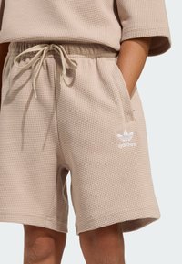 Béžové šortky z textúrovanej pleteniny s elastickým pásom, šnúrkou, bočnými vreckami a bielym logom adidas na ľavej nohe.