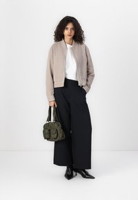 Béžová kordurová bomber bunda, biele tričko, široké čierne nohavice a tmavozelená kabelka. Modelka stojí so vzpriamenou postúrou na bielom pozadí.