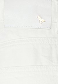 Una tela blanca con una textura suave presenta una etiqueta cosida y un pequeño acento de abeja dorada. El diseño es minimalista y limpio.
