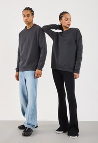 To personer står mot en ensfarget bakgrunn, iført mørkegrå hettegensere; den ene har på seg lyseblå jeans og svarte sko, mens den andre har svarte bukser og joggesko.