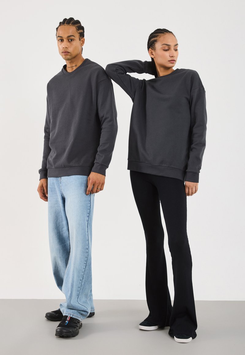 To personer står mot en ensfarget bakgrunn, iført mørkegrå hettegensere; den ene har på seg lyseblå jeans og svarte sko, mens den andre har svarte bukser og joggesko.