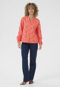 Blusa corallo con bottoni, ricamata con fiori, maniche lunghe a sbuffo e scollatura a V, abbinata a jeans blu scuro a gamba dritta e sandali marroni.