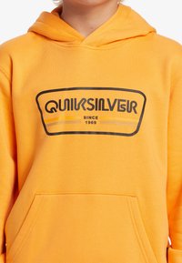Žlutá mikina s kapucí vyrobená z bavlněné směsi, s černým logem Quiksilver a textem "od roku 1969", s klokánkovou kapsou na přední straně.