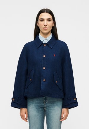 Femme aux cheveux raides et foncés portant une veste bleu marine boutonnée sur une chemise bleu clair à col et un jean délavé clair, faisant face.