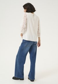 White blouse with lace three-quarter sleeves, back keyhole detail, paired with loose blue denim pants and black boots. Plain background.