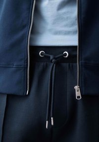 Giacca blu navy con zip, indossata sopra una camicia azzurra chiara; presenta una cerniera argentata, vita elasticizzata e cordino con occhielli argentati.