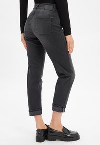 Femme portant un jeans noir coupé aux chevilles avec des revers et des mocassins noirs, vue de dos et de la taille vers le bas sur fond blanc.