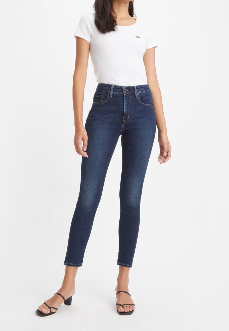 Mörkblå högmidjade skinny jeans av denim, med klassisk femficksdesign och subtil blekning på låren. Kombinerade med en vit t-shirt.