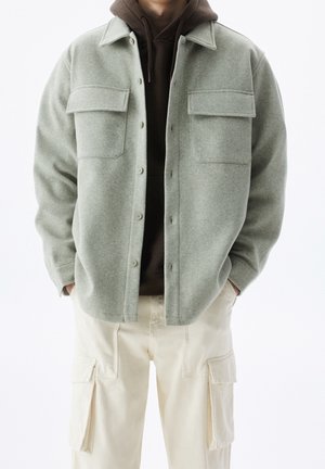 Homme portant une veste en laine gris clair boutonnée sur un hoodie marron foncé et un pantalon cargo crème, les mains dans les poches.