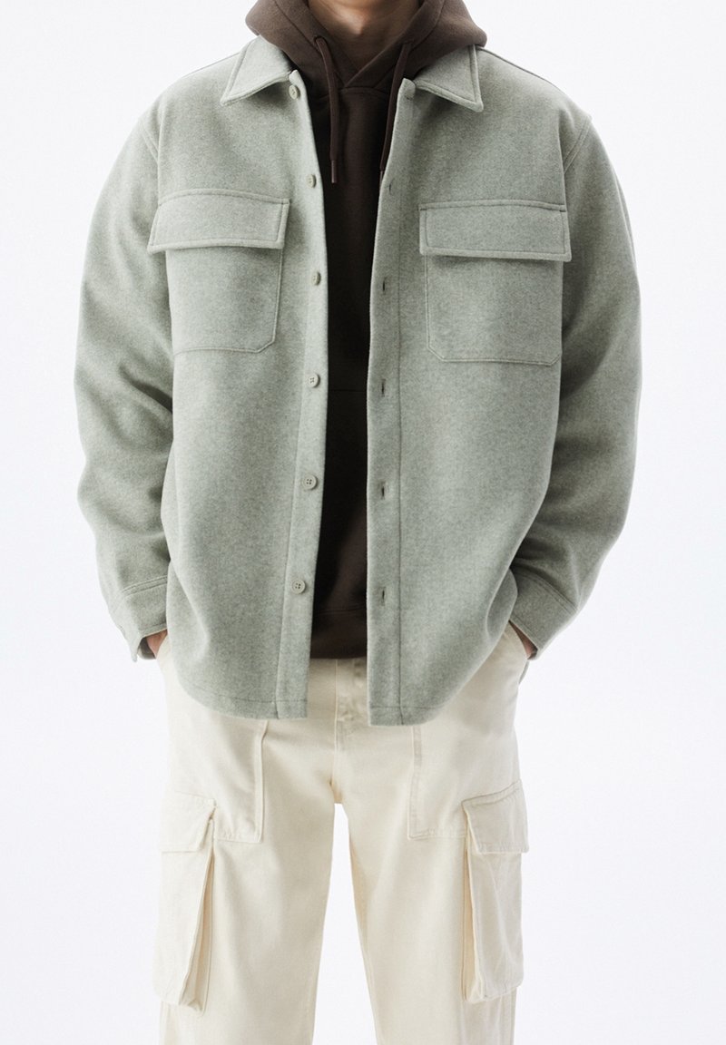 Homme portant une veste en laine gris clair boutonnée sur un hoodie marron foncé et un pantalon cargo crème, les mains dans les poches.
