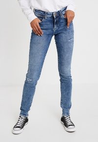 Persona que lleva unos jeans ajustados azules con un ligero desgaste, una camiseta blanca de manga larga y zapatillas negras de caña alta, de pie con las manos cerca de los bolsillos.