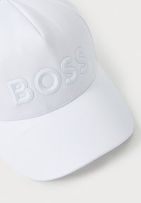 Hvit caps laget av teksturert stoff med en hevet, blank "BOSS"-logo over forsiden. Buet skygge, elegant design.