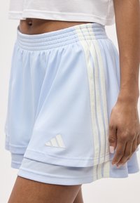 Κοντινό πλάνο ατόμου που φοράει ανοιχτό μπλε στρωμένα σορτς Adidas με λευκές ρίγες και λογότυπο, συνδυασμένα με λευκό κοντό τοπ.