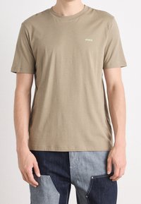 Mand iført en ensfarvet olivengrøn Hugo T-shirt og patchwork denim jeans med mørkeblå og lyseblå paneler, står med armene afslappet.